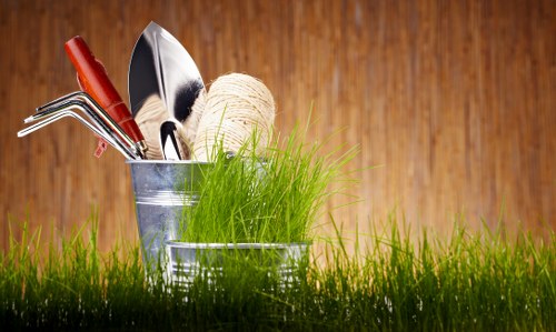 The image shows a close-up view of a small patch of a garden or outdoor space, featuring a grassy area in the foreground with lush green grass blades. Behind the grass, there is a collection of gardening tools and materials arranged in a metal container, including a pair of pruning shears with red handles, a small garden trowel, a metal rake, and a ball of twine. The background consists of warm, natural wooden surface, possibly fencing or siding, suggesting an outdoor garden setting. The overall scene emphasizes tools and natural elements associated with gardening and outdoor maintenance, with natural light illuminating the scene, enhancing the vibrant green of the grass and the metallic shine of the garden tools.