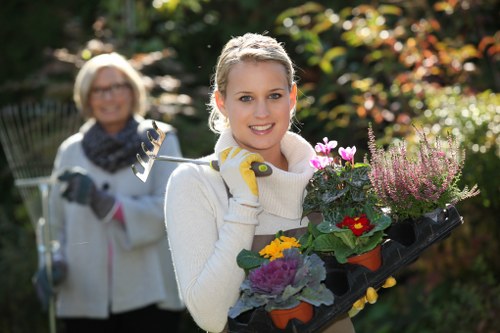 A young woman with blonde hair, wearing a white sweater and gardening gloves, is holding a black tray filled with various colorful potted plants and flowers, including pink, yellow, and purple blooms, in an outdoor garden setting. Behind her, an older woman with glasses, a white jacket, and a gardening fork in hand is slightly out of focus, smiling and standing within a lush garden with green foliage, shrubs, and trees. The garden appears well-tended, with a paved pathway and natural sunlight filtering through the leaves, suggesting a peaceful, landscaped outdoor space typical of a suburban garden area in Anerley, South London. This setting is relevant for gardening services such as hedge trimming, lawn care, and garden maintenance, emphasizing the care and vibrant growth of outdoor greenery.